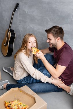 Neşeli adam kız arkadaşını karton kutuların yanında pizzayla besliyor.