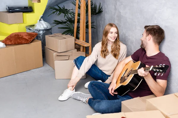 高兴的女人看着男朋友在盒子边弹奏吉他 搬迁的概念 — 图库照片
