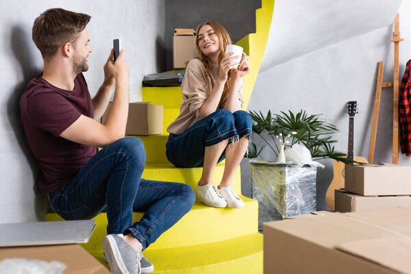 joyful man taking photo of girlfriend with cup while sitting on stairs near boxes