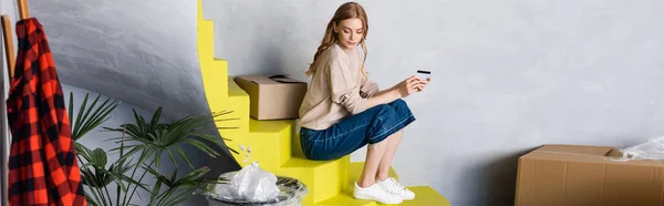 クレジットカードを持って箱の近くの階段に座っている若い女性の水平方向のイメージ — ストック写真