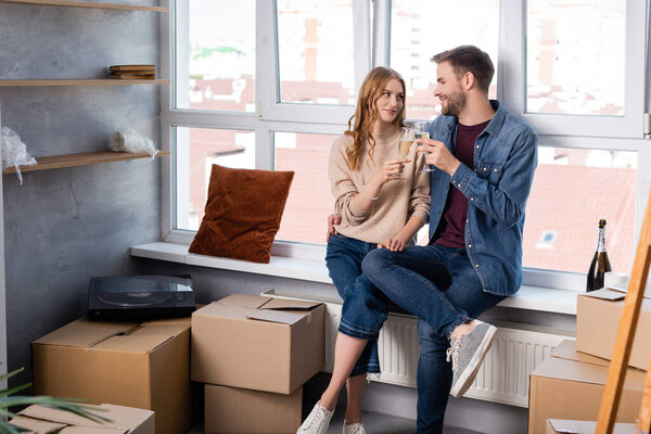 pleased man hugging girlfriend and clinking glasses with champagne near carton boxes, moving concept 