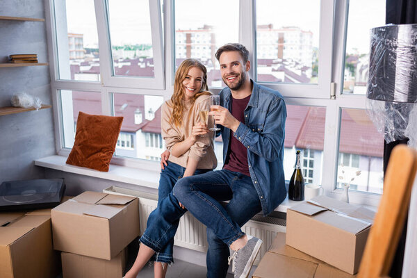 selective focus of pleased couple toasting glasses with champagne near carton boxes, moving concept 