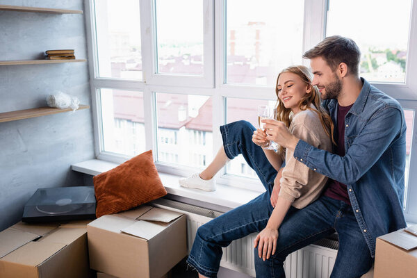 pleased couple toasting glasses with champagne near windows and carton boxes, moving concept 