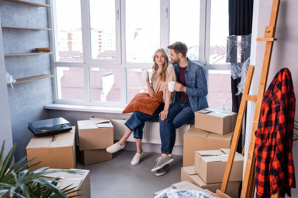 pleased couple holding cups of tea near carton boxes, moving concept 