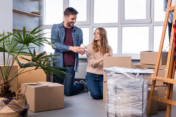 man and woman looking at cup while unpacking carton boxes in new home, moving concept