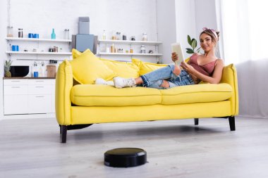 Dijital tabletle kanepede uzanmış bir kadın ve yerde de robotik elektrikli süpürge.
