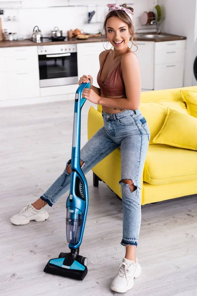Housewife holding vacuum cleaner and sitting on yellow couch at home
