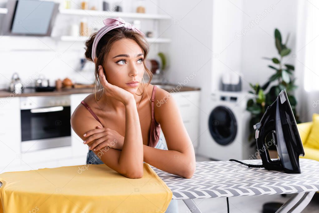 Dreamy woman in headband looking away near t-shirt on ironing board at home 