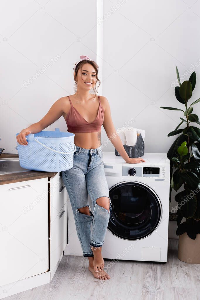 Young housewife with laundry basket standing near washing machine at home 