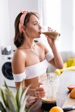 Blender 'ın yanında smoothie ve evde meyve içen genç bir kadının seçici odak noktası 