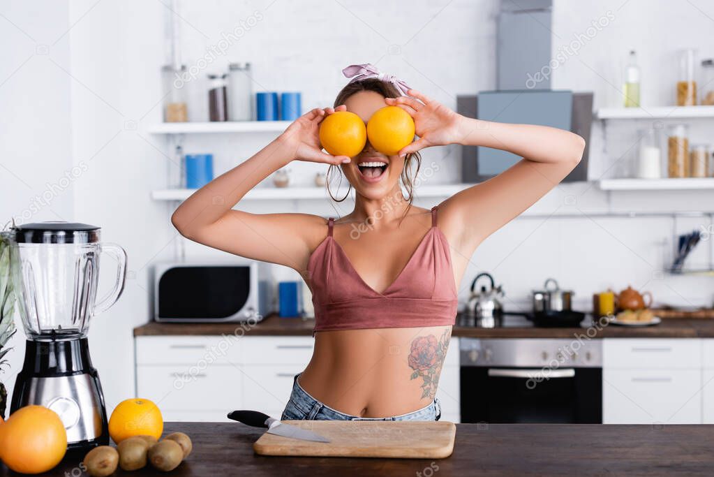 Selective focus of tattooed woman holding oranges near eyes beside cutting board and blender 