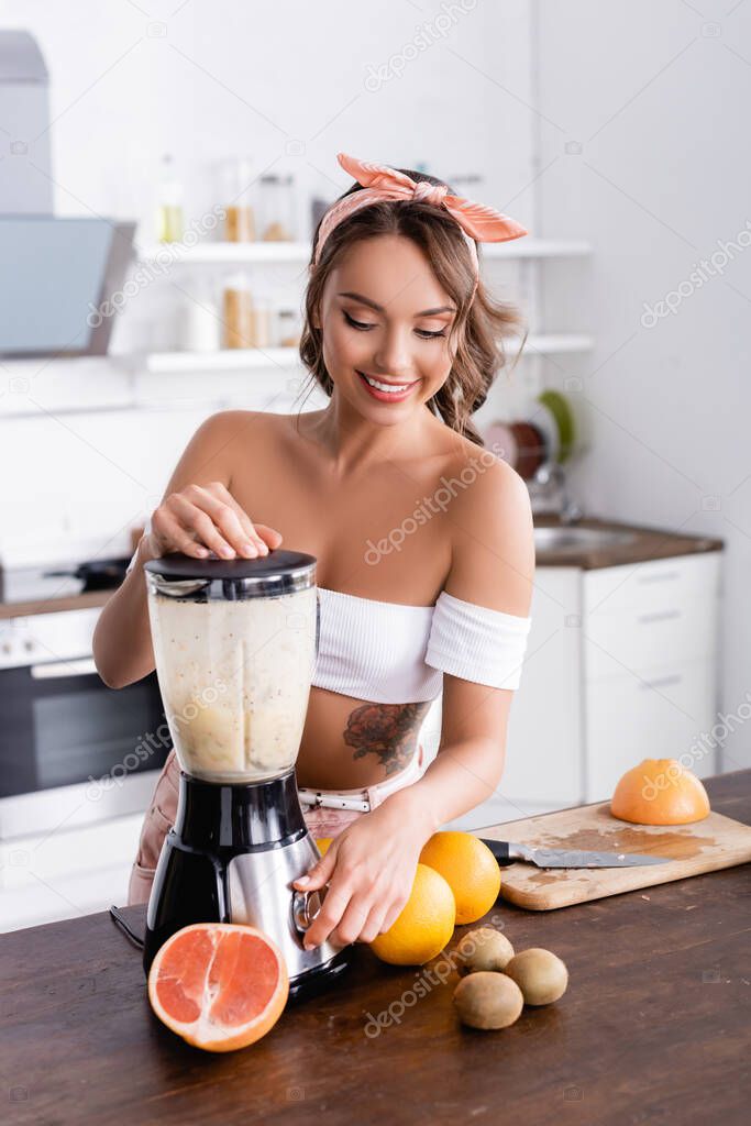 Selective focus of young woman preparing smoothie near ripe fruits on kitchen table 