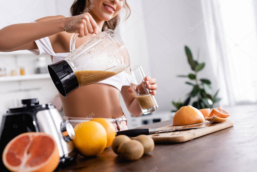 Cropped view of young woman pouring smoothie in glass near organic fruits on kitchen table 