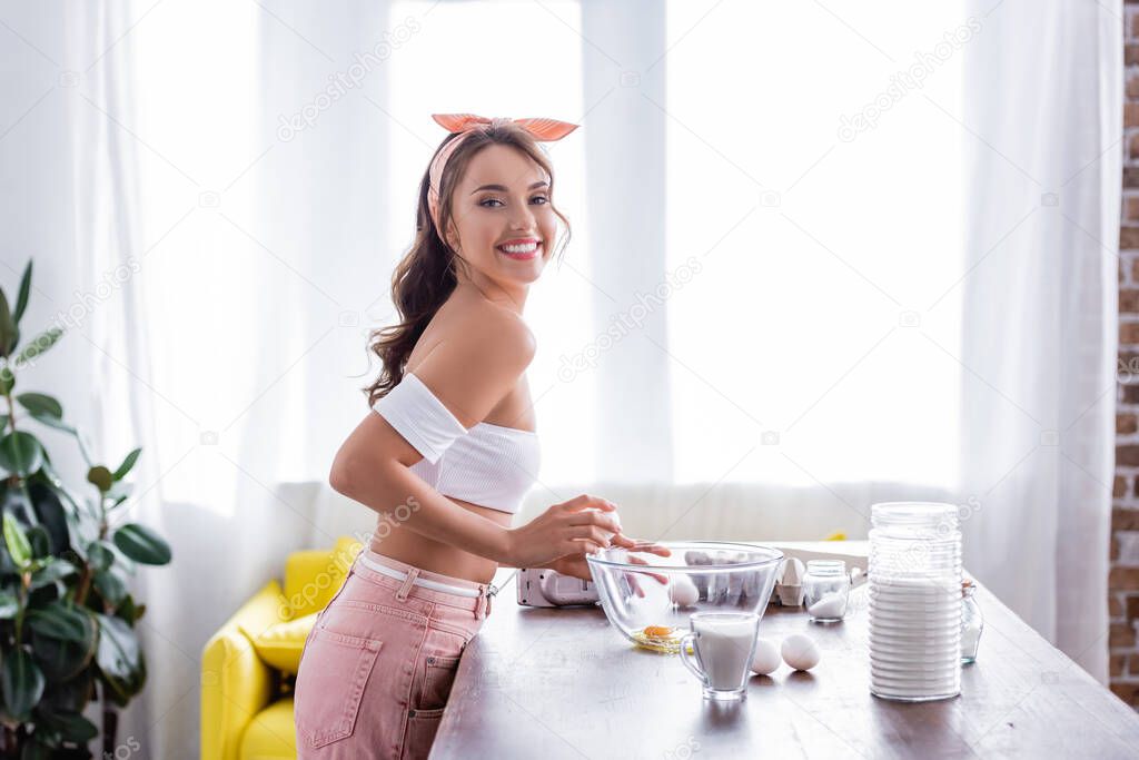 Side view of young woman looking at camera while breaking eggs in bowl in kitchen 