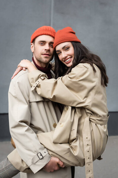 stylish man holding in arms woman in beanie hat and trench coat 