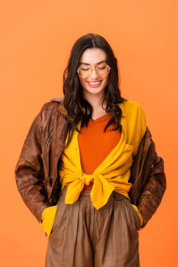 young woman in autumn outfit standing with hands in pockets and looking down isolated on orange 