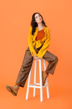 full length of young woman in autumn outfit and boots sitting on white stool on orange  clipart