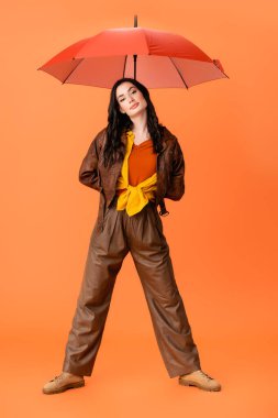 full length of young fashionable woman in autumn outfit and boots standing with umbrella on orange clipart