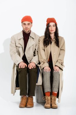 young brunette woman in trench coat sitting near man in beanie hat on white clipart