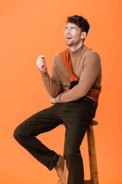 excited man in autumn outfit and glasses sitting on wooden stool and laughing on orange 