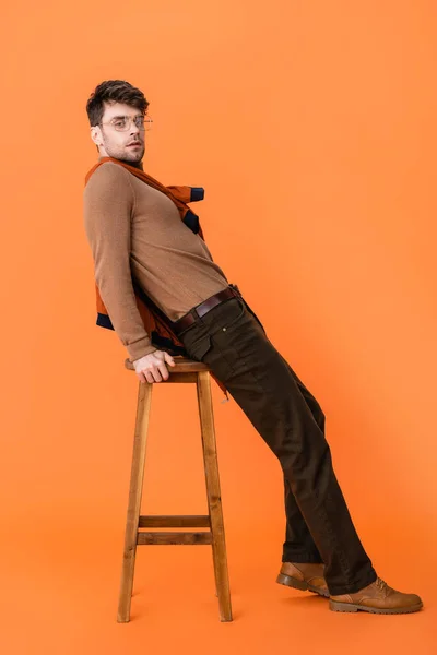 stylish man in autumn outfit and glasses leaning on wooden stool on orange