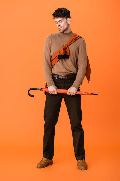 full length of stylish man in autumn outfit holding umbrella on orange 