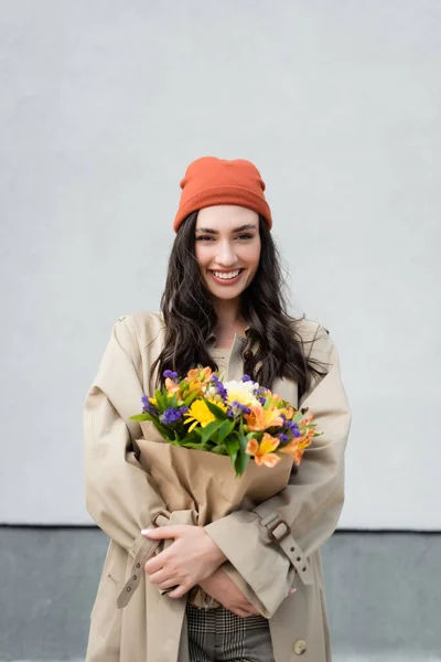 ビーニー帽子とトレンチコートのスタイリッシュな女性は 灰色の壁の近くに花の花束を保持 — ストック写真