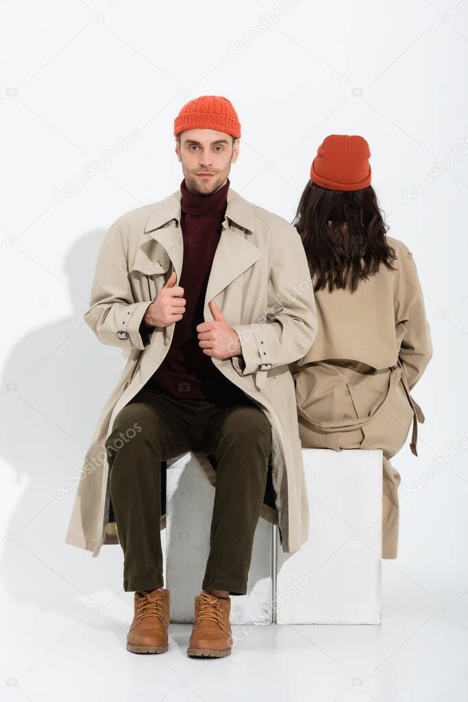stylish man in beanie hat touching trench coat near woman sitting on white