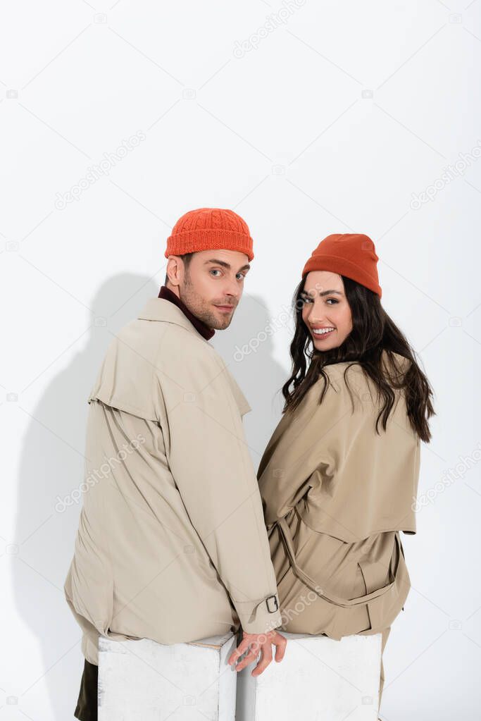 trendy couple in beanie hats and trench coats sitting and looking at camera on white 