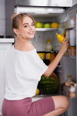 smiling woman in white t-shirt taking mustard from fridge and looking at camera