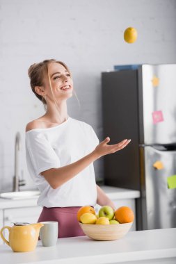 Çaydanlık ve fincanın yanında dikilirken olgun limonla hokkabazlık yapan neşeli genç kadın.