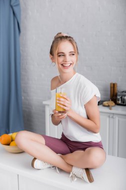 Mutfak masasında bacakları çapraz oturan ve elinde portakal suyu tutan beyaz tişörtlü mutlu genç kadın.