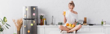 horizontal concept of young blonde woman sitting on kitchen table with fresh fruits and orange juice clipart