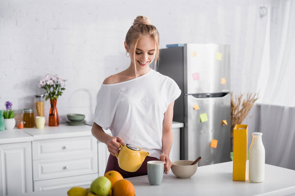 joyful woman in white t-shit pouring tea into cup near bowl and fresh fruits