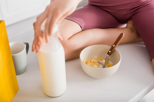 обрезанный вид молодой женщины, открывающей бутылку молока, сидя на столе рядом с миской с кукурузными хлопьями