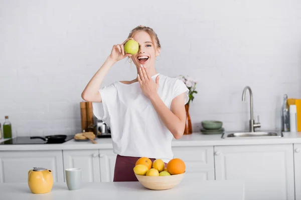 興奮ブロンド女性タッチあごで目をカバーしながら新鮮なリンゴでキッチン — ストック写真