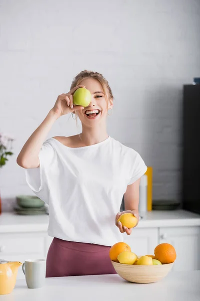Веселая Молодая Женщина Закрывает Глаза Яблоком Держа Лимон Кухне — стоковое фото