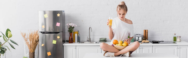 horizontal concept of young blonde woman sitting on kitchen table with fresh fruits and orange juice
