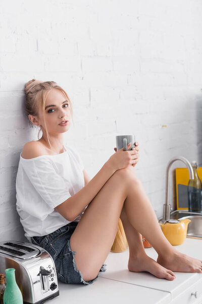 sensual barefoot woman looking at camera while sitting on kitchen table with cup of tea
