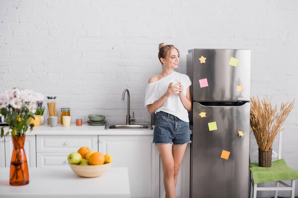 selective focus of young blonde woman in white t-shirt and shorts holding cup while standing in kitchen