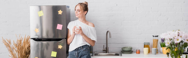 panoramic shot of young woman standing near fridge in kitchen with cup of tea and looking away