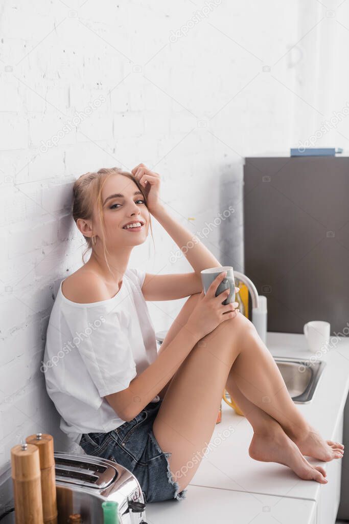 young barefoot woman in white t-shirt and shorts sitting on kitchen table with cup of tea 