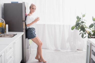 barefoot blonde woman standing near refrigerator with cup of tea clipart