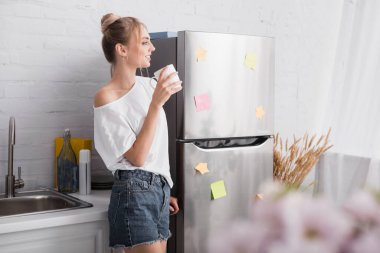 selective focus of blonde woman in white t-shirt and denim shorts standing in kitchen with cup of tea clipart