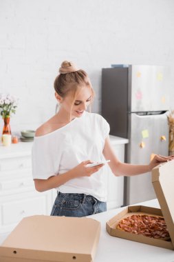Mutfakta pizzayla kutuyu açarken beyaz tişörtlü genç bir kadın cep telefonuyla mesajlaşıyor.