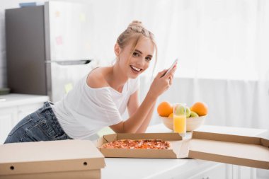 Heyecanlı genç bir kadın pizzanın yanında akıllı telefondan sohbet ediyor ve mutfakta taze meyve yiyor.