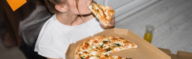 Mutfakta yerde otururken pizza yiyen genç bir kadının panoramik görüntüsü.