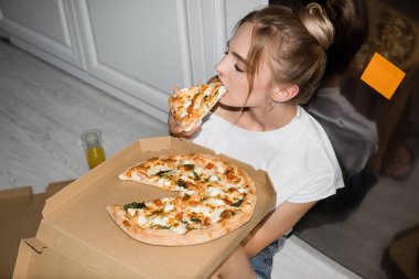 Yüksek açılı genç sarışın kadın mutfakta yerde otururken pizza yiyor. 