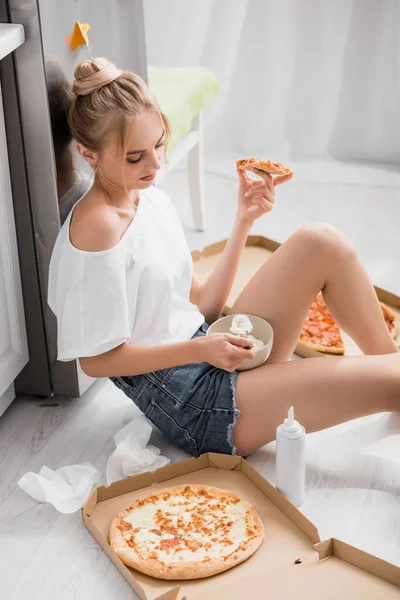 身穿白色T恤和短裤的年轻女子坐在厨房的地板上 手里拿着一碗奶油和披萨 — 图库照片