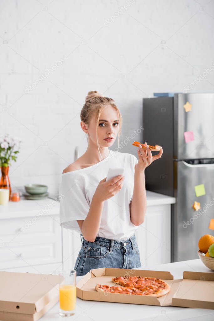 blonde woman holding smartphone and pizza while looking at camera in kitchen
